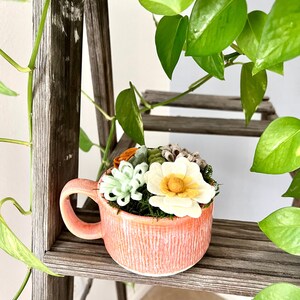 Op de afbeelding: Een koraalkleurige keramische mok gevuld met kunstmatige vetplanten en bloemen. De mok heeft een textuur en een oor. De opstelling bevat witte, groene en oranje bloemelementen, tegen een achtergrond van groen mos.