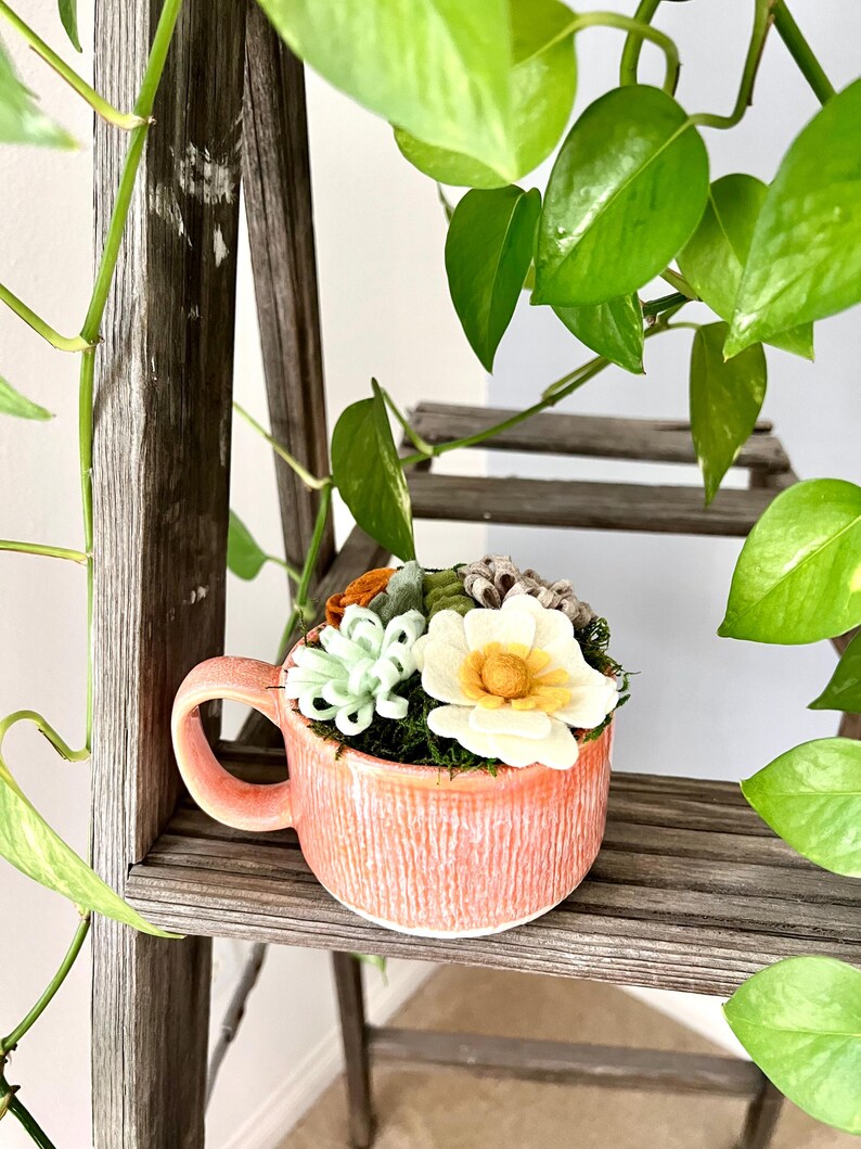 Op de afbeelding: Een koraalkleurige keramische mok gevuld met kunstmatige vetplanten en bloemen. De mok heeft een textuur en een oor. De opstelling bevat witte, groene en oranje bloemelementen, tegen een achtergrond van groen mos.