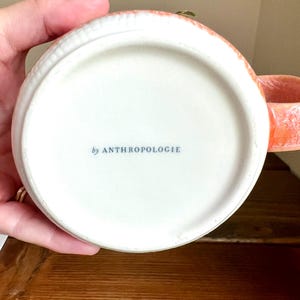 Op de afbeelding: Een witte keramische mok met een koraalkleurige handgreep en rand. De onderkant van de mok heeft de tekst "by ANTHROPOLOGIE" in blauw. De mok staat op een houten oppervlak.