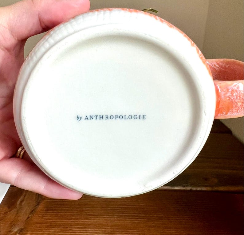 Op de afbeelding: Een witte keramische mok met een koraalkleurige handgreep en rand. De onderkant van de mok heeft de tekst "by ANTHROPOLOGIE" in blauw. De mok staat op een houten oppervlak.