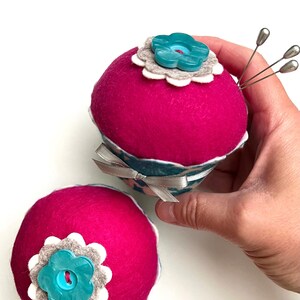 May include: Two handmade pincushions. The pincushions are a vibrant fuchsia color, with a flower-shaped button and a gray and white felt flower embellishment. One pincushion has silver pins inserted. A teal and white patterned ribbon wraps around the base.