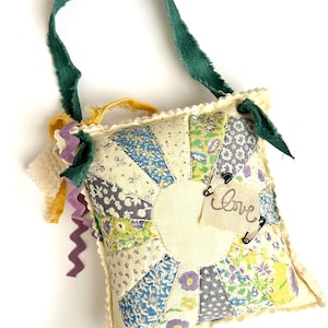 May include: A small, square, patchwork pillow with a sunburst design in shades of blue, yellow, and gray. The pillow has a green ribbon handle and a small tag that says "love". Decorative ribbons and trim are attached to the pillow.