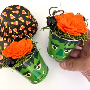 May include: Two green metal buckets with a Frankenstein face design, each holding an orange felt flower and green moss. A stack of black and orange candy corn cupcake liners is in the background. The buckets have small silver handles.