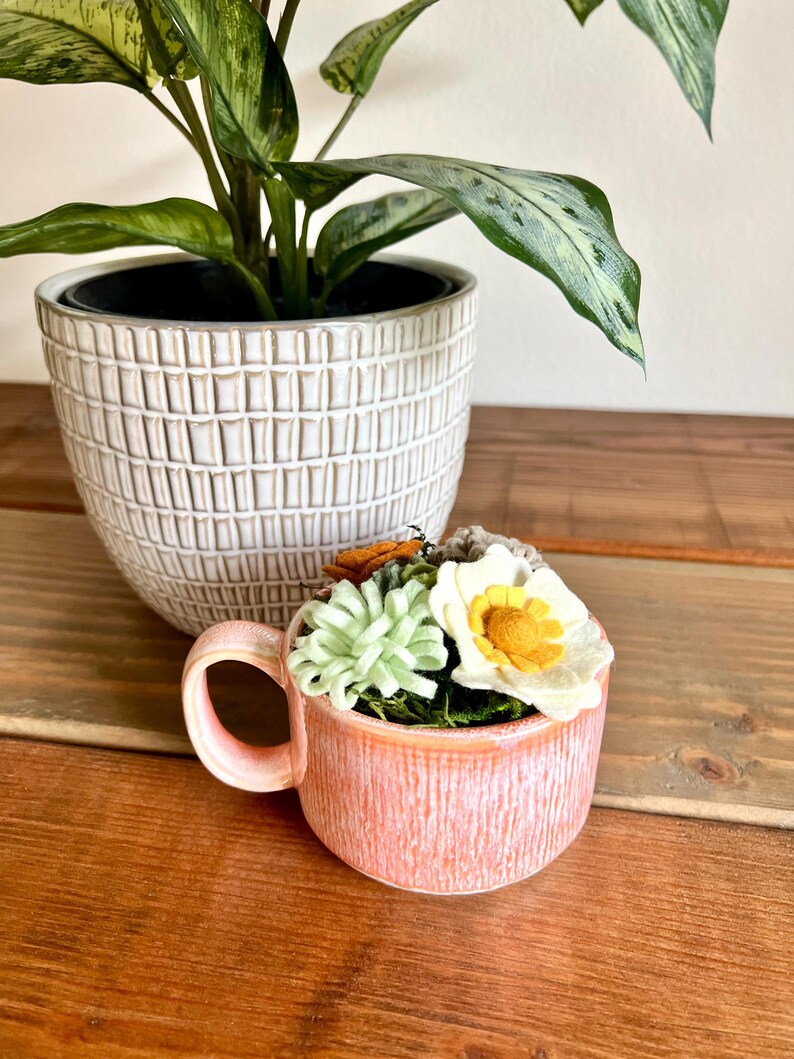 Op de afbeelding: Een kleine, perzikroze keramische mok gevuld met kunstbloemen en groen. De mok heeft een gestructureerd oppervlak en een gebogen handvat. Een grotere witte plantenbak met een rasterpatroon en een groene plant staat op de achtergrond.