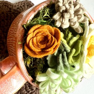 Op de afbeelding: Een handgemaakte perzikkleurige theekop gevuld met viltbloemen in tinten oranje, groen, cr&egrave;me en bruin. De theekop staat op een bruine gehaakte onderzetter. De bloemen zijn zo gerangschikt dat ze op een miniatuurtuin lijken.