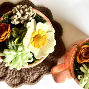 Op de afbeelding: Twee perzikroze theekopjes gevuld met viltbloemen en groen. E&eacute;n kopje heeft een grote witte bloem met een gele kern, en de andere heeft een oranje roos. De kopjes staan op een bruine gehaakte onderzetter.