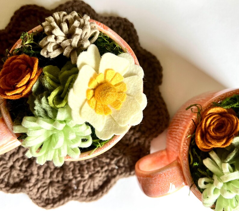 Op de afbeelding: Twee perzikroze theekopjes gevuld met viltbloemen en groen. E&eacute;n kopje heeft een grote witte bloem met een gele kern, en de andere heeft een oranje roos. De kopjes staan op een bruine gehaakte onderzetter.