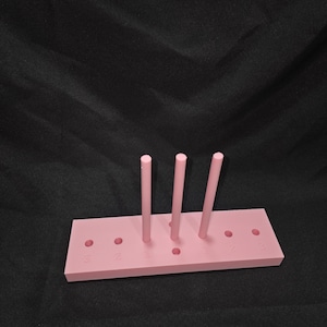 May include: A pink bow maker tool with three vertical dowels and numbered holes. The rectangular base has six holes and is made of a solid pink material. The background is a solid black.