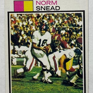 May include: A vintage football card featuring Norm Snead, quarterback for the Giants, in action. The card has a white border with pink and yellow accents. The image shows Snead in a white uniform with the number 16, holding a football.