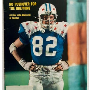 May include: Sports Illustrated magazine cover featuring football player John Matuszak in a blue jersey with the number 82. The cover text reads "NO PUSHOVER FOR THE DOLPHINS". The player wears a white helmet with blue stripes and red stars.