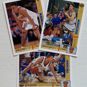 May include: Three vintage basketball trading cards featuring New York Knicks players. The cards display action shots of John Starks, Charles Oakley, and Mark Jackson in their Knicks uniforms. The cards have a wooden border and the Knicks logo.
