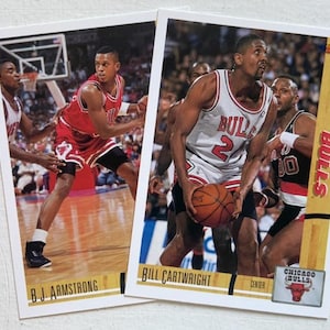 May include: Two basketball trading cards featuring Chicago Bulls players. One card shows B.J. Armstrong in a red uniform. The other card shows Bill Cartwright in a white and red uniform, holding a basketball. The cards have the Bulls logo.