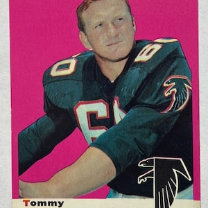 May include: A vintage football card featuring Tommy Nobis of the Atlanta Falcons. The card has a pink background and displays Nobis in his team jersey. The Falcons logo is visible, along with the text "Tommy Nobis" and "Linebacker."