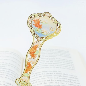 May include: Gold-colored bookmark with an ornate design featuring koi fish and lotus flowers. The top section has a detailed illustration of koi fish swimming in a pond. The handle also has koi fish and lotus flower designs.