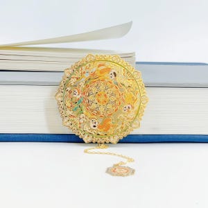 May include: A gold metal bookmark with an elaborate circular design, incorporating orange, green, and white details. The bookmark includes a decorative chain and a small, ornate tag. It rests on a stack of books.