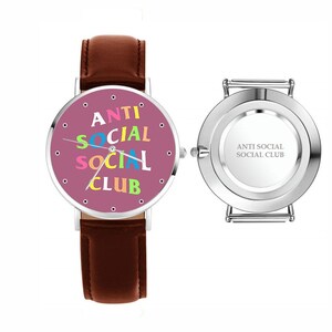 Puede incluir: Reloj de pulsera con correa de cuero marr&oacute;n y caja plateada. La esfera malva muestra el texto "ANTI SOCIAL SOCIAL CLUB" en colores vivos. La parte trasera del reloj tambi&eacute;n tiene el texto "ANTI SOCIAL SOCIAL CLUB".