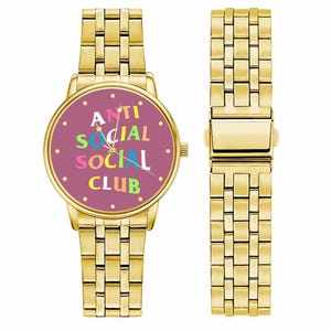 Puede incluir: Reloj de pulsera dorado con esfera morada que muestra el texto "ANTI SOCIAL SOCIAL CLUB" en letras coloridas. El reloj tiene una correa dorada a juego. Se muestra junto a una correa de reloj separada.