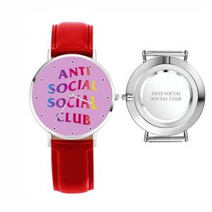 May include: A wristwatch with a red leather band and a round face. The watch face is light purple with the words "ANTI SOCIAL SOCIAL CLUB" in colorful text. The back of the watch is silver with the same text.