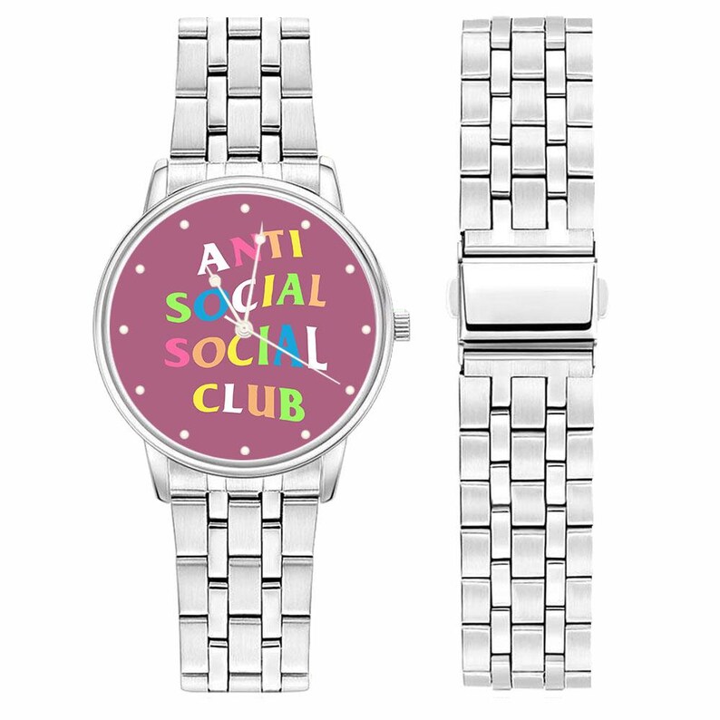 Puede incluir: Reloj de pulsera plateado con una correa de eslabones rectangulares. La esfera del reloj es de color rosa intenso con las palabras "ANTI SOCIAL CLUB" en letras de colores. El reloj se muestra junto a una correa suelta.