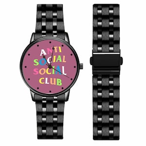 Puede incluir: Reloj de pulsera negro con esfera redonda y correa a juego. La esfera del reloj es de color rosa polvoriento y muestra las palabras "ANTI SOCIAL CLUB" en letras de colores.