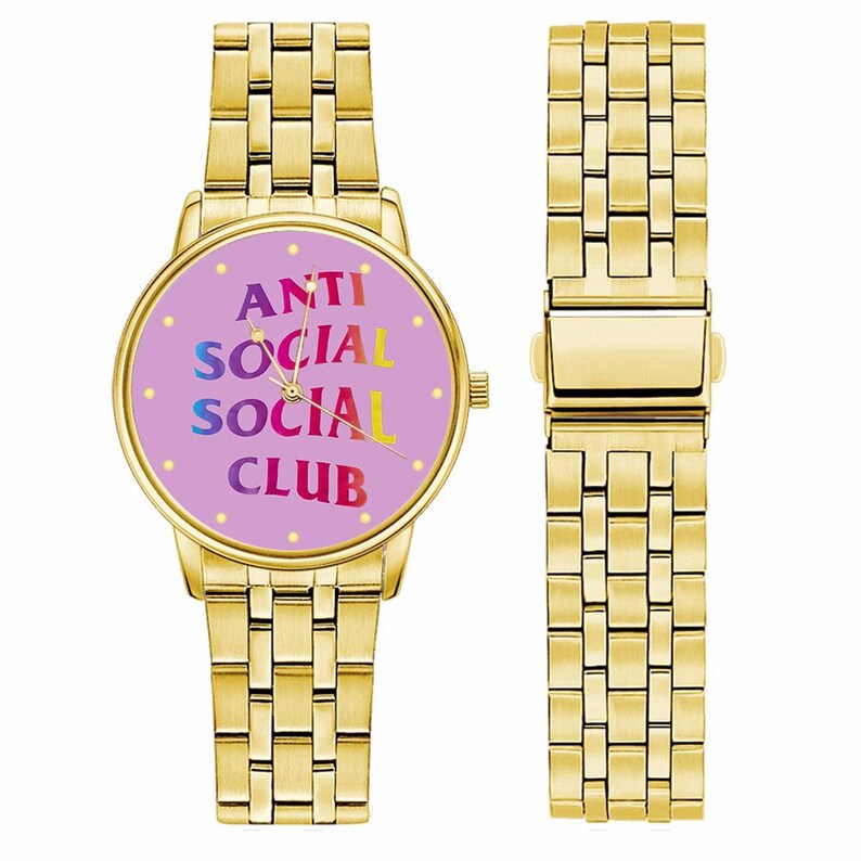 May include: Gold-toned wristwatch with a rectangular link band. The watch face is lavender with the words "ANTI SOCIAL SOCIAL CLUB" in rainbow colors. The watch is shown next to a detached band.