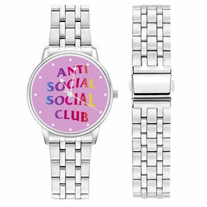May include: A silver-toned wristwatch with a light purple face. The watch face features the text "ANTI SOCIAL SOCIAL CLUB" in rainbow colors. The watch band is made of rectangular links. A separate watch band section is also visible.