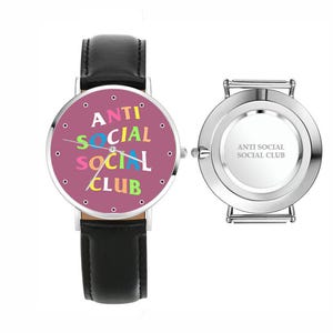 Puede incluir: Reloj de pulsera con correa de cuero negro y esfera redonda. La esfera rosa muestra el texto "ANTI SOCIAL SOCIAL CLUB" en letras coloridas. La parte trasera plateada del reloj tambi&eacute;n tiene el mismo texto.
