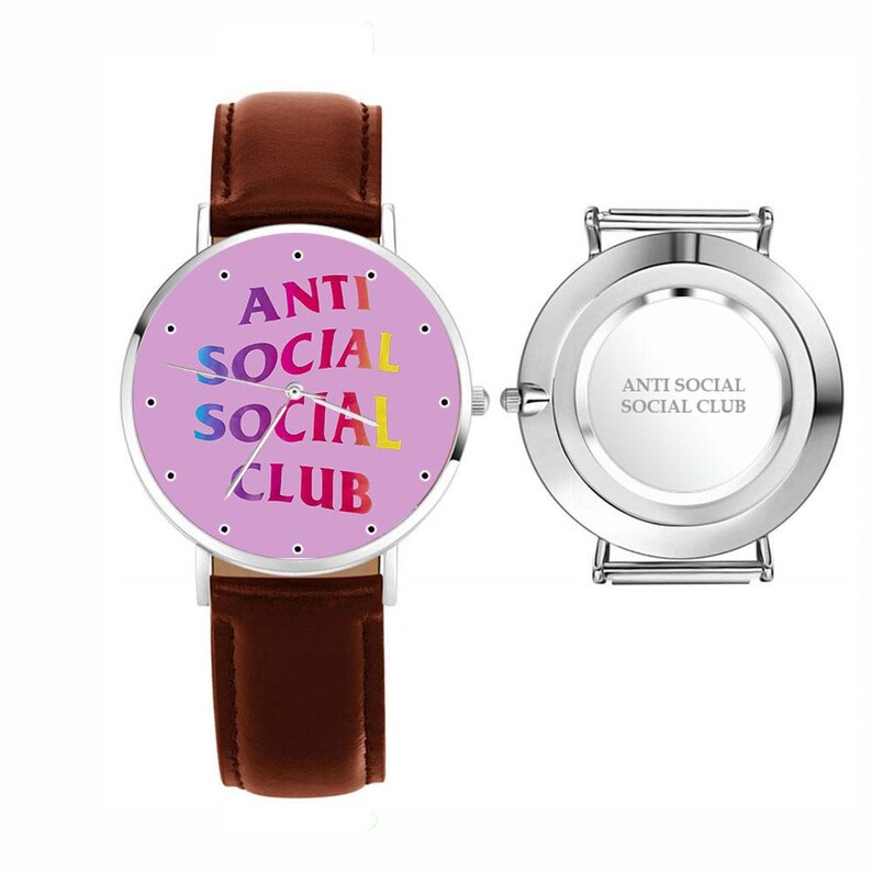 May include: A wristwatch with a brown leather band and a round face. The watch face is light purple with the words "ANTI SOCIAL SOCIAL CLUB" in colorful text. The back of the watch is silver with the same text.