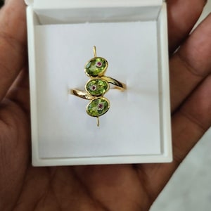 May include: A gold ring featuring three oval-shaped green gemstones, each accented with a small red stone in the center. The ring is presented in a white box, showcasing its elegant design and craftsmanship.