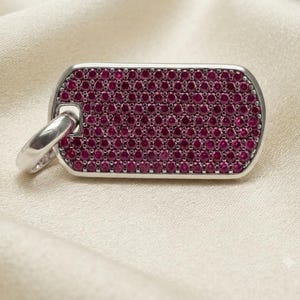 May include: A rectangular dog tag pendant featuring a polished silver-tone metal frame filled with a dense grid of small, round, deep pinkish-red stones. A smooth, thick silver-tone metal jump ring is attached to the top left corner of the pendant.