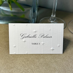 Può includere: Un segnaposto rettangolare bianco con linee verticali e motivi a stella in rilievo. Il testo "Gabriella Palmer" è stampato in un carattere corsivo nero, con "TABLE 1" stampato in un carattere nero sans-serif più piccolo e in grassetto.