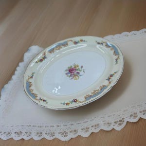 May include: Oval porcelain serving platter with a cream-colored background and a floral design in the center. The rim features blue and gold accents with floral patterns. The platter is placed on a white lace doily.