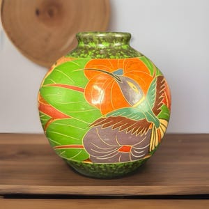 May include: A round, hand-painted ceramic vase with a green base and rim. The vase features a vibrant design of a hummingbird in flight, set against a backdrop of orange flowers and green leaves. The artwork is detailed and colorful.
