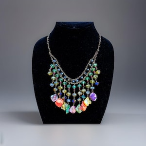 May include: A multi-strand necklace with a silver chain and colorful beads. The necklace features green, blue, and yellow beads, with larger, irregularly shaped, iridescent shell pendants in shades of orange, pink, and purple. The necklace is displayed on a black mannequin.