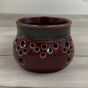 May include: A handcrafted ceramic vessel with a deep burgundy glaze. The pot features a dark green band near the rim and is adorned with a pattern of circular cutouts. This decorative piece is suitable for various uses.