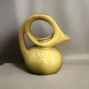 May include: A yellow ceramic pitcher with a unique, curved handle and a rounded base. The spout is angled, and the pitcher has a glossy finish. The design is modern and sculptural, suitable for decorative use.