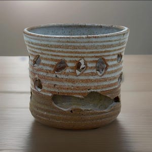 May include: Handmade ceramic vessel with a cylindrical shape. The pottery features horizontal bands of tan and white, with irregular cut-out shapes. The base is a darker tan color, and the rim is a light blue-gray. A unique, rustic home decor item.