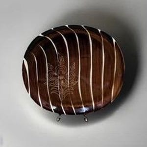 May include: A brown, oval-shaped decorative plate with white vertical stripes and a floral design in the center. The plate is displayed on a metal stand against a neutral background.