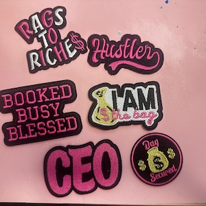May include: A collection of embroidered patches with motivational phrases in pink and black. Phrases include "Rags to Riches," "Hustler," "Booked Busy Blessed," "CEO," "I AM the bag," and "Bag Secured."