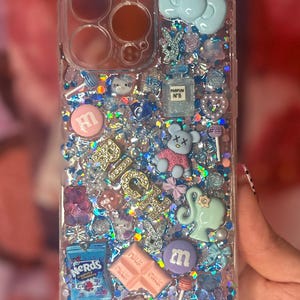 May include: Clear phone case with a glittery, multi-colored design. Adorned with various charms, including a perfume bottle, a teddy bear, a bow, and candy. The word "RICH" is spelled out in gold letters.