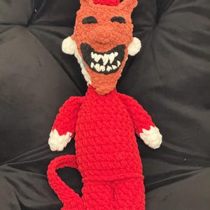 May include: A handmade crochet devil doll with a smiling face. The doll features a red body, arms, and tail, with white accents on the ears and hands. The face is orange with black eyes and a wide, toothy grin. The doll is a unique, whimsical creation.