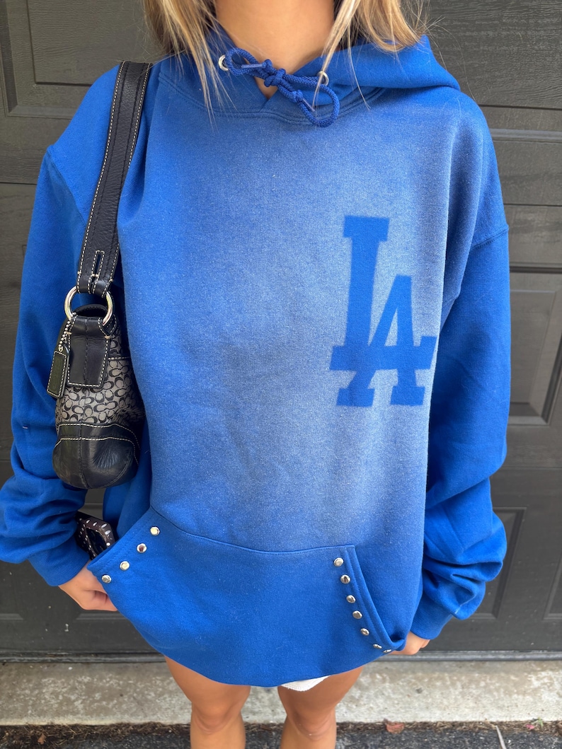 Puede incluir: Una sudadera con capucha azul real con las letras "LA" en azul m&aacute;s claro en la parte delantera. La sudadera tiene un bolsillo de canguro con tachuelas plateadas. Un bolso negro con correa de cadena es visible a la izquierda.