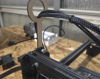 May include: A close-up of a laser engraving machine with a wooden component featuring a circular handle. The machine has a black frame with measurement markings and a black cable. The background is a light-colored wooden surface.