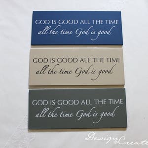 Op de afbeelding: Drie houten borden met de tekst "GOD IS GOOD ALL THE TIME all the time God is good" in witte en zwarte letters. De borden zijn geschilderd in blauw, beige en grijs.