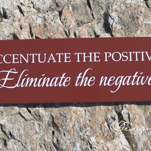 Op de afbeelding: Een rood houten bord met witte tekst die luidt "Accentueer het positieve, elimineer het negatieve".