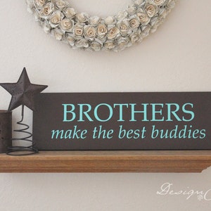 Op de afbeelding: Een bruin houten bord met de tekst "BROTHERS make the best buddies" in turquoise blauwe letters. Het bord staat op een houten plank met een metalen ster en een kaarsenhouder.