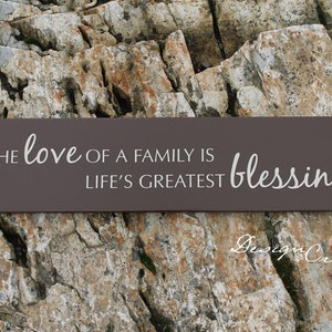May include: A brown wooden sign with white text that reads "The love of a family is life's greatest blessing".