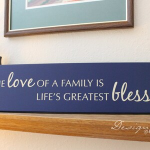 May include: A navy blue wooden sign with white lettering that reads "The love of a family is life's greatest blessing".