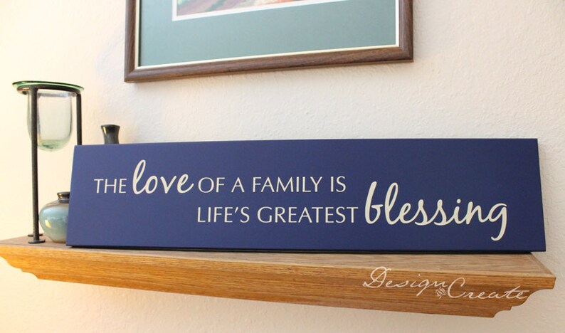 May include: A navy blue wooden sign with white lettering that reads "The love of a family is life's greatest blessing".