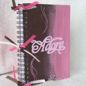 May include: A brown and pink spiral bound notebook with the word "Adore" written in pink on the front cover. The notebook has brown and pink ribbons tied around the spine.